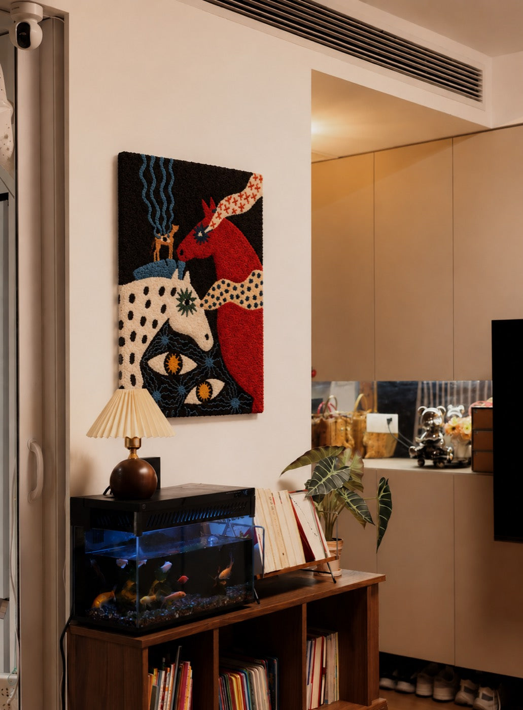 A hand-tufted wool panel of two folk-art horses in red and cream on black ground, hung above a pleated lamp and bookshelf