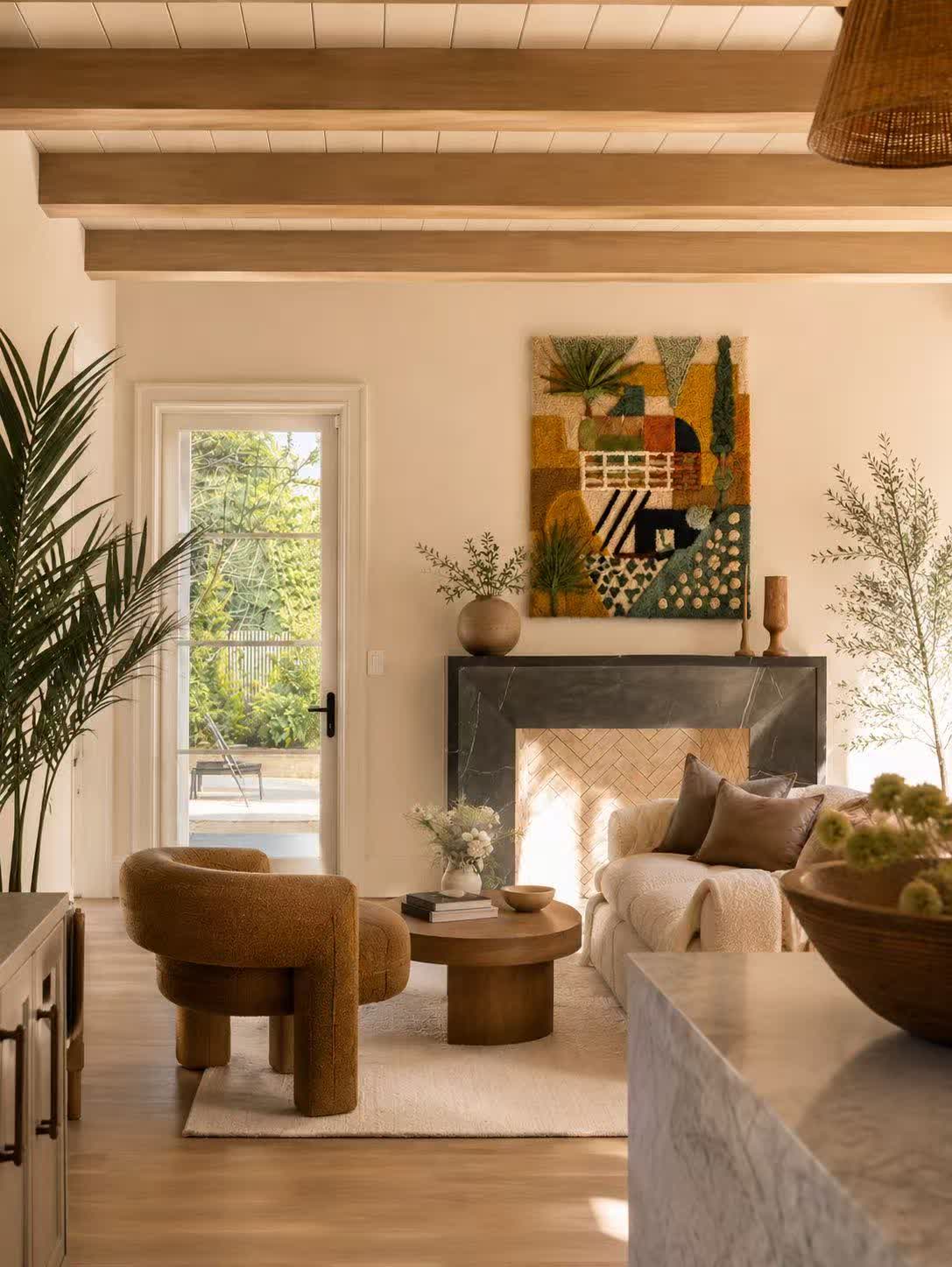 A hand-tufted wool tapestry in ochre, olive and teal hung above a stone fireplace in a sunlit beamed living room