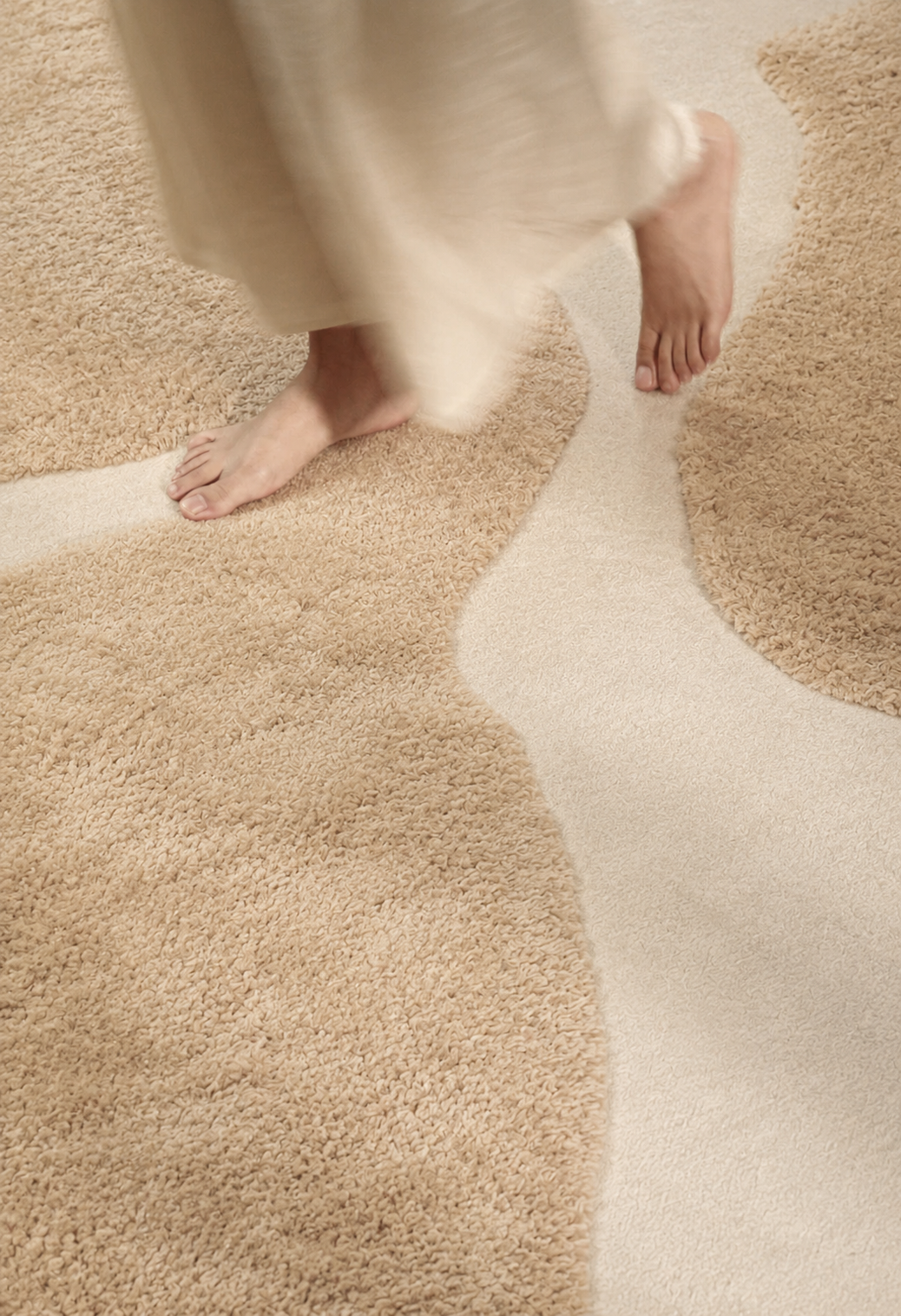 Hewn Earth Studio Bloom rug in sand and cream with a soft, textured design, shown on a wooden floor with feet.