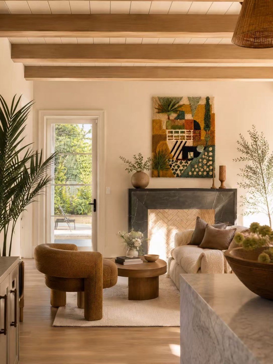 A hand-tufted wool tapestry in ochre, olive and teal hung above a stone fireplace in a sunlit beamed living room