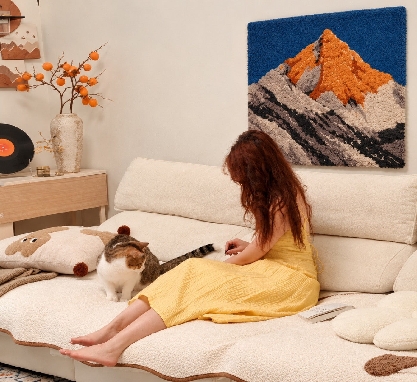 A hand-tufted wool mountain landscape in cobalt and rust hung above a cream sofa, with a cat and a reader below it