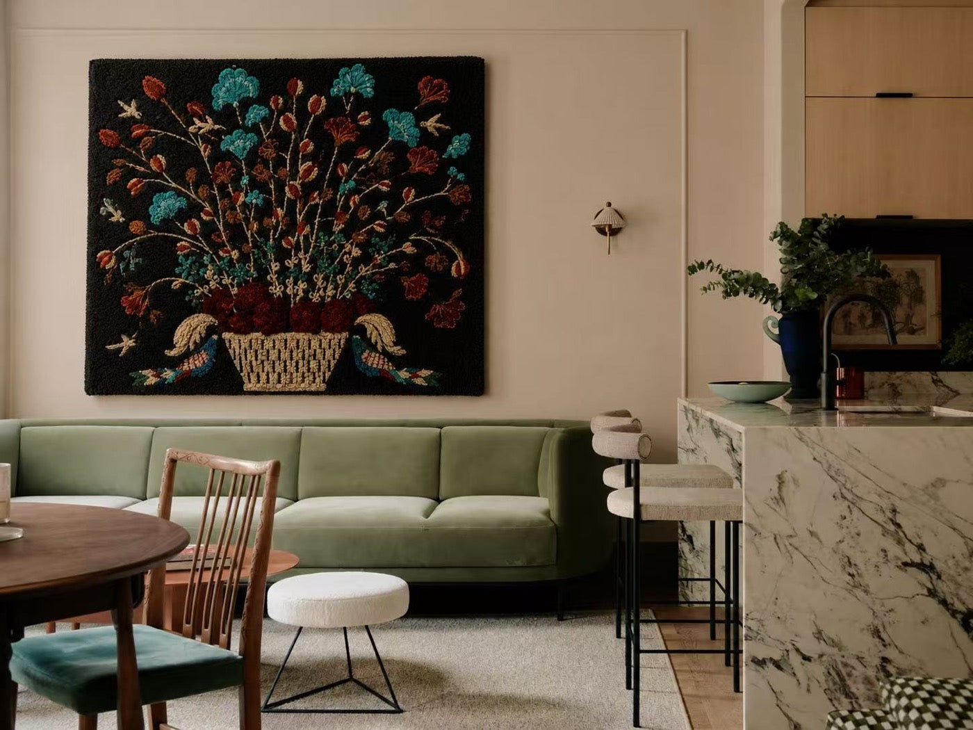 A hand-tufted wool still life of a flower basket on black ground hung above a sage green sofa in a marble kitchen-dining room