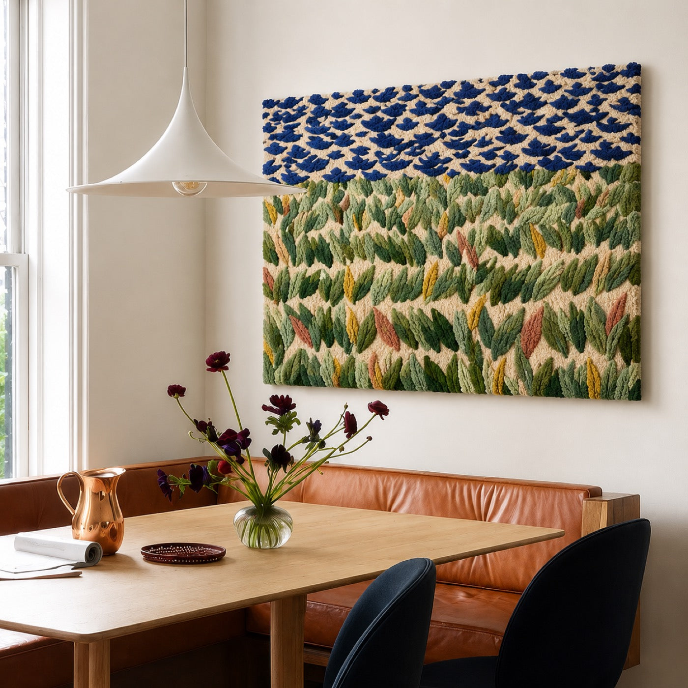 A hand-tufted wool meadow panel of cobalt blossoms above green leaves hung over a leather banquette in a sunlit breakfast nook