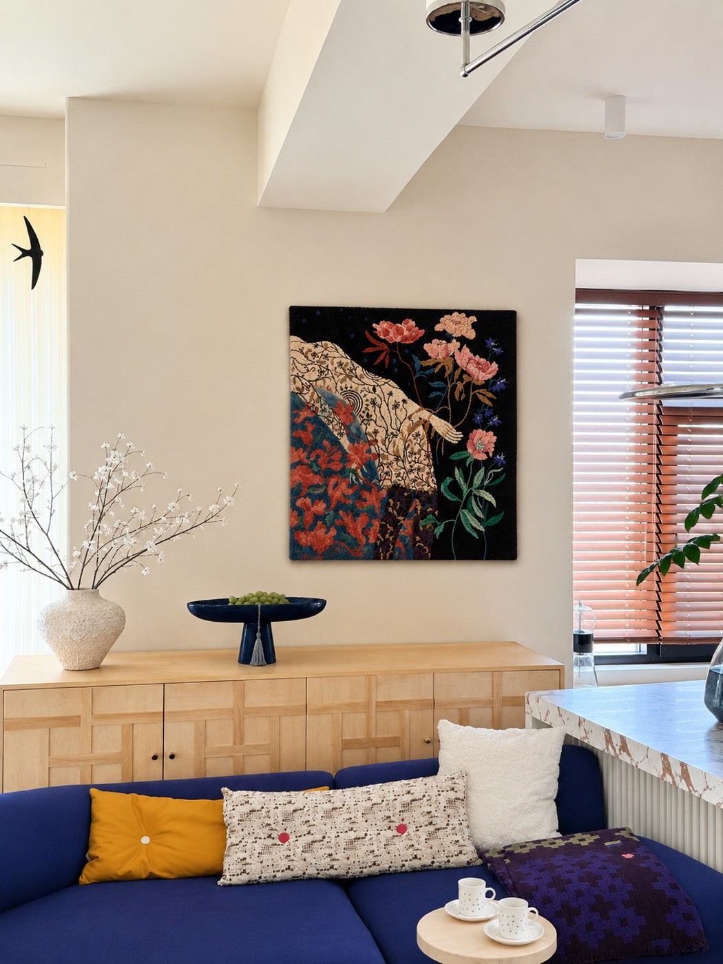 A hand-tufted wool panel of a hand reaching through peonies on black ground, hung above a blue sofa and oak sideboard