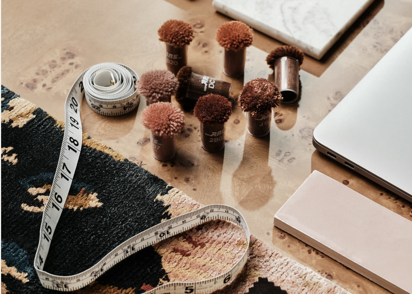 A flatlay of wool colour tufts, a tape measure, stone samples and a laptop on a designer's desk